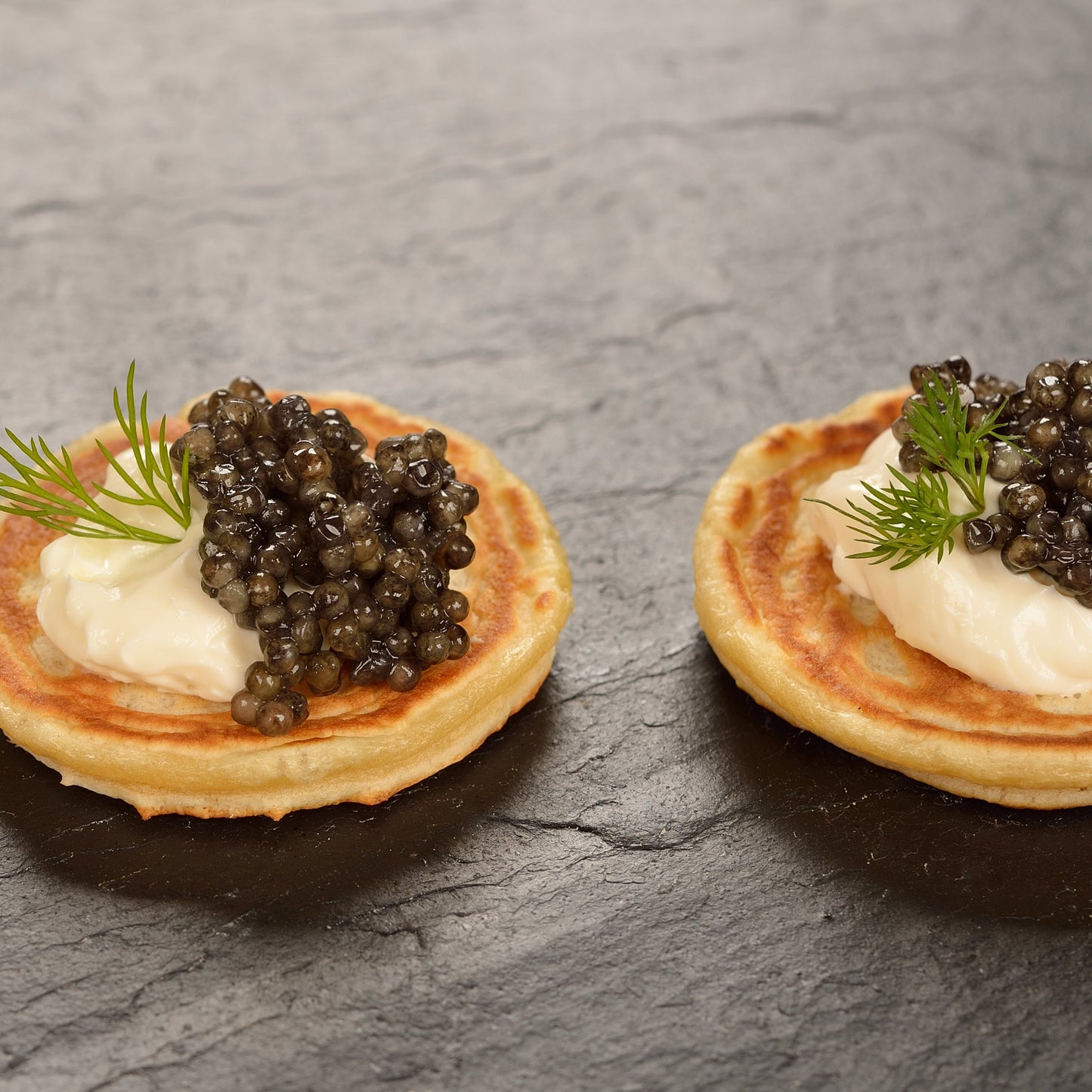 Three small blinis topped with cream and caviar, each garnished with a sprig of dill, arranged on a dark slate surface.