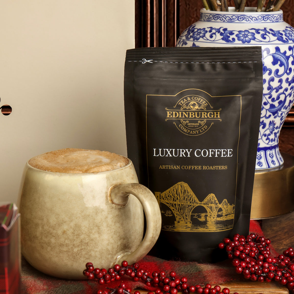 A bag of Edinburgh Luxury Coffee stands next to a beige ceramic mug filled with coffee, with red berry decorations and a blue-and-white vase in the background.