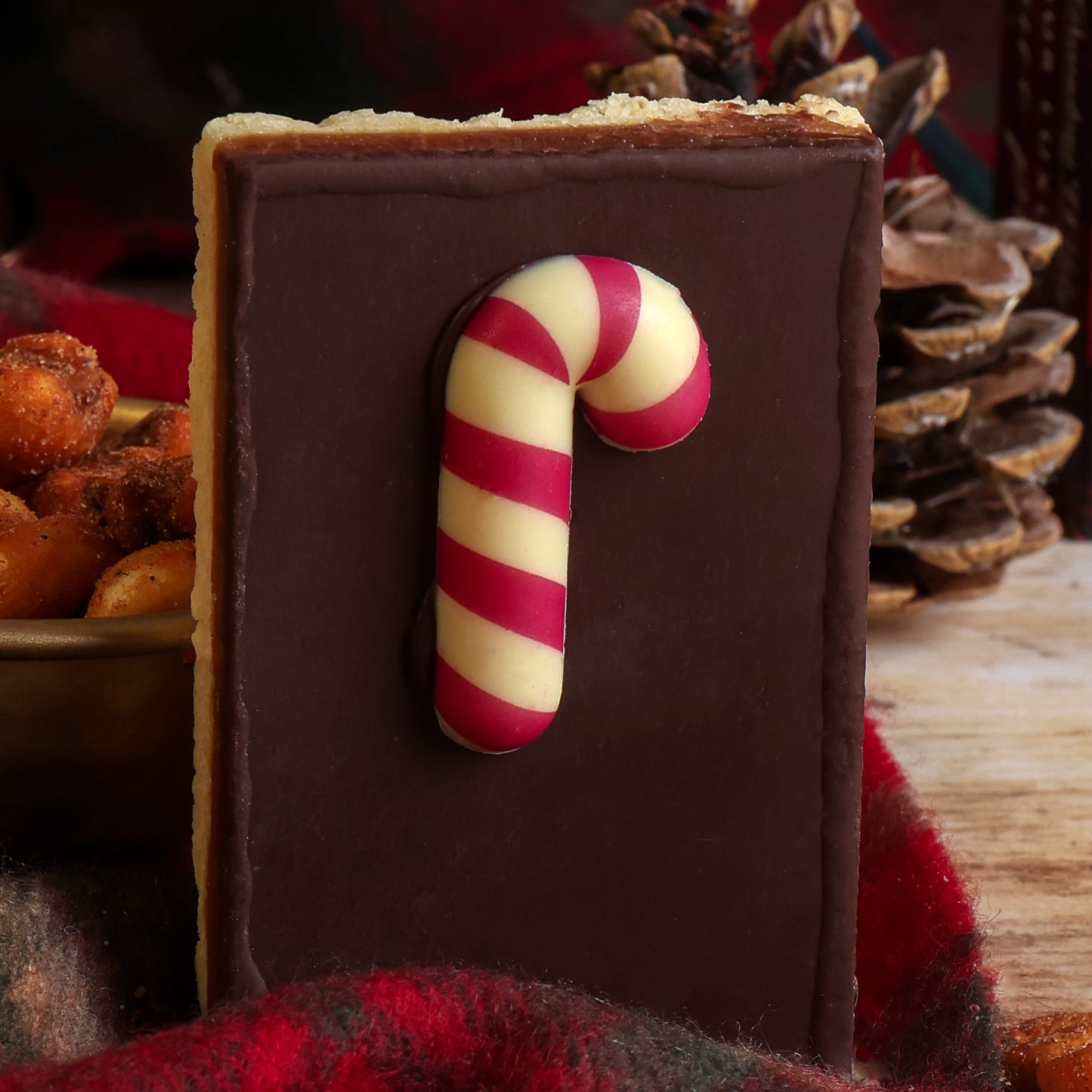 A rectangular flapjack topped with chocolate and a candy cane decoration in red and white stripes, set against a rustic Christmas background with pinecones and red fabric.
