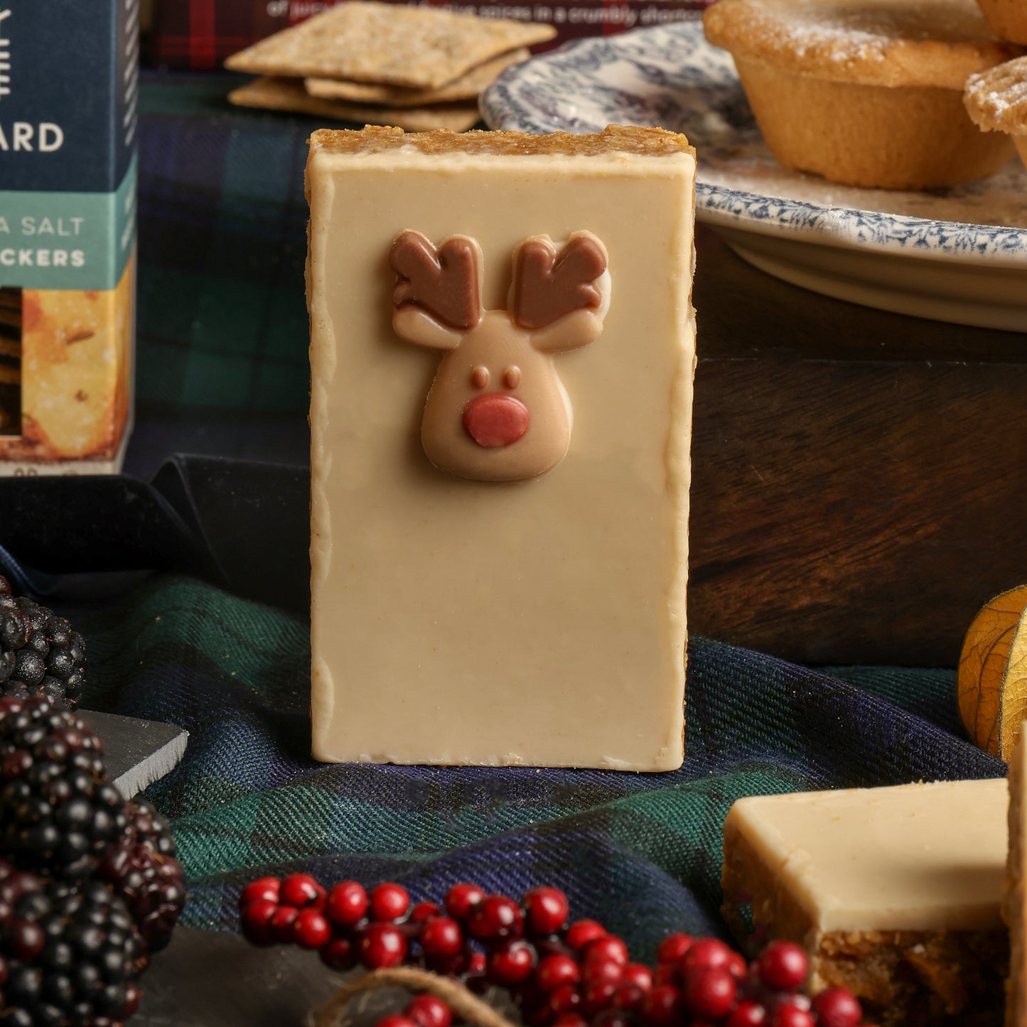 A rectangular white chocolate flapjack slice with a small reindeer face decoration on the front, styled on a dark plaid cloth with Christmas decorations and mince pies around it.