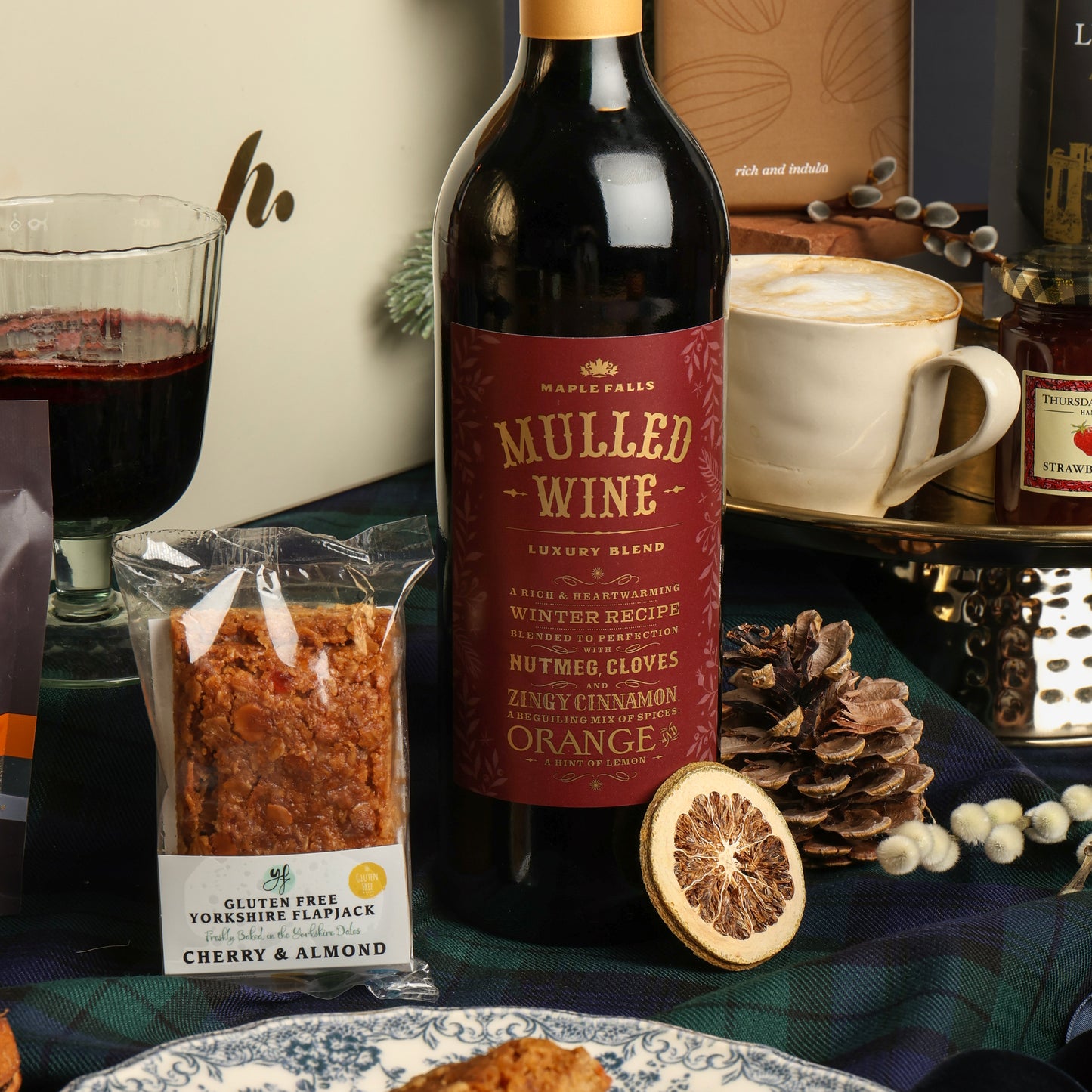 A bottle of Maple Falls Mulled Wine is centred on a festive table setting with a glass of mulled wine, a mug of coffee, a packaged cherry and almond flapjack, a dried orange slice, pinecones, and a plaid tablecloth.