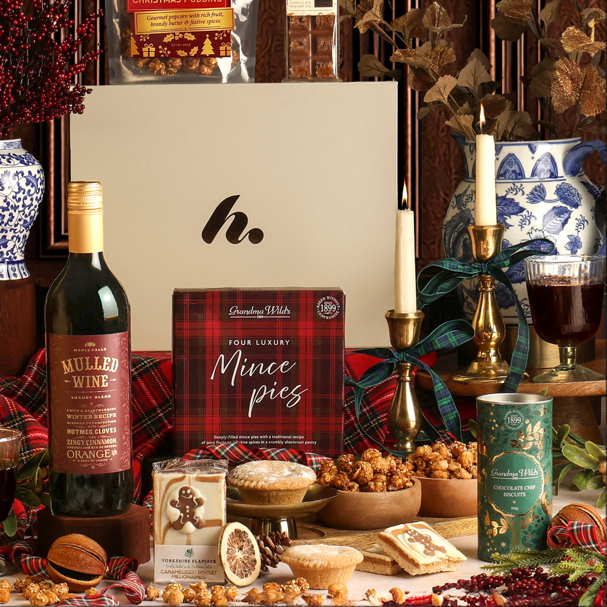 The Festive Night In Christmas hamper with its contents displayed, including a bottle of mulled wine, a box of mince pies, a tin of biscuits and popcorn, styled with dried fruit, candles and plaid fabric in a Christmas setting.