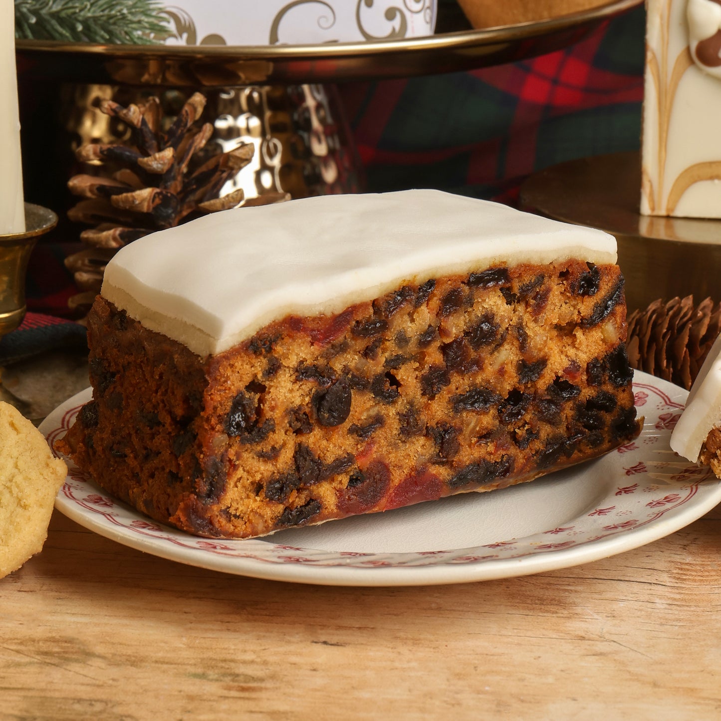 A Christmas fruit cake with white icing on top, served on a white plate with a red floral border, set on a wooden table with Christmas decorations in the background.