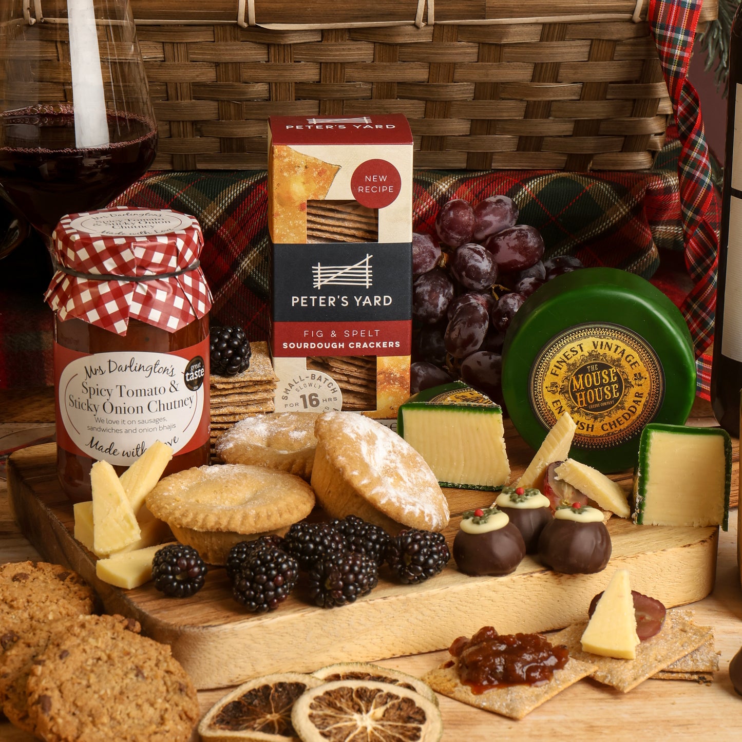 A collection of food and wine from the Classic Christmas Food and Wine Hamper, arranged on a wooden serving board, featuring a jar of Spicy Tomato & Sticky Onion Chutney, a box of Peter's Yard Sourdough Crackers, a green wheel of The Mouse House Cheddar cheese, cheese wedges, chocolates, a glass of red wine, with blackberries and dried orange slices to decorate the scene.