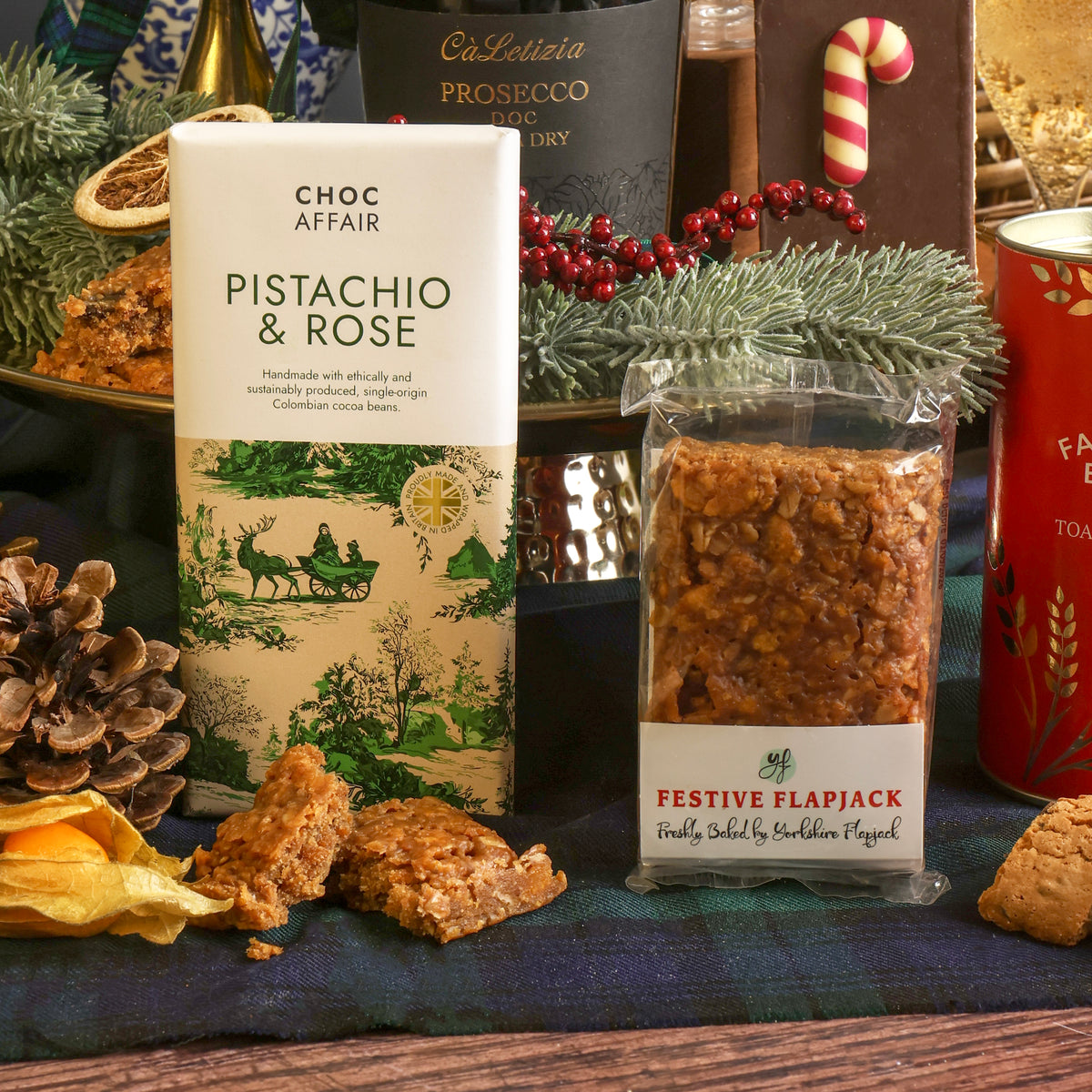 A festive product display featuring a bar of Choc Affair Pistachio & Rose chocolate alongside a clear packet of festive flapjack, surrounded by Christmas decorations including pine branches, pinecones, red berries, and a candy cane flapjack.