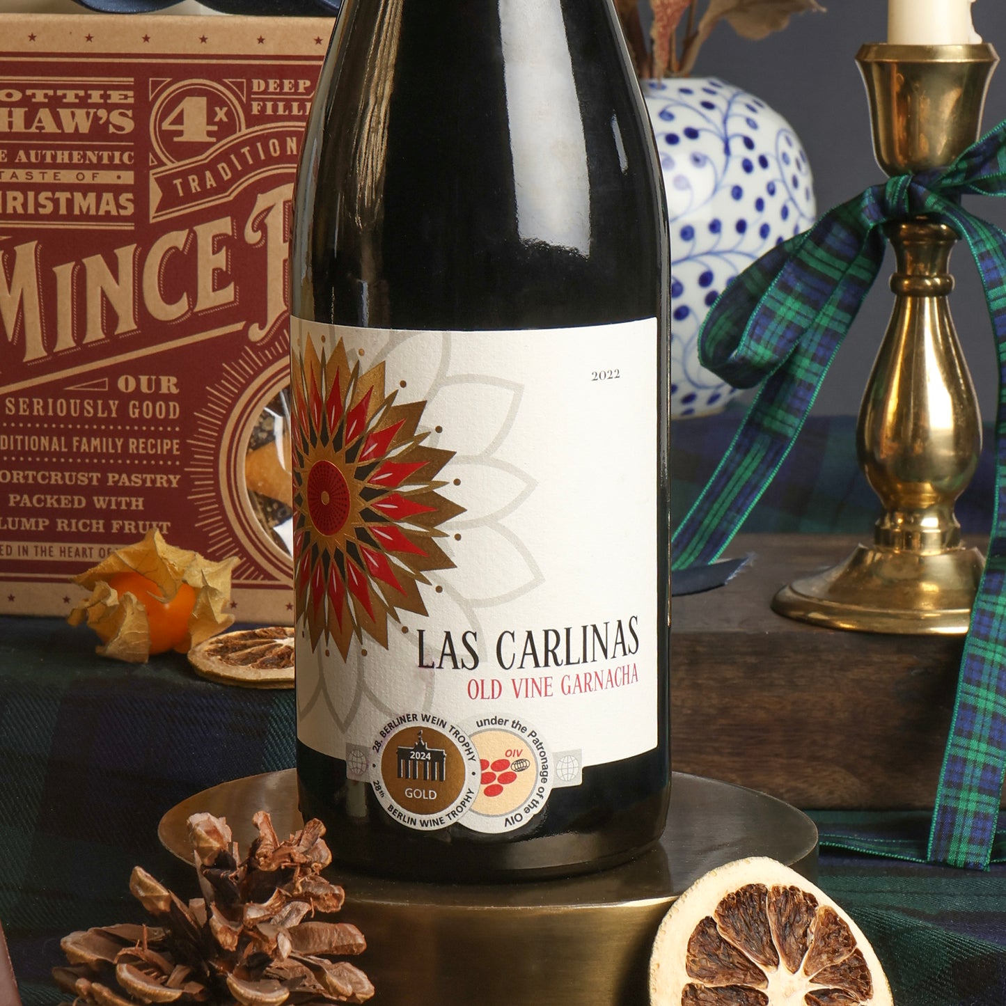 A bottle of Las Carlinas red wine displayed on a brass pedestal with Christmas-themed props including sliced orange, a pinecone, a candlestick tied with ribbon, and a box of mince pies in the background.