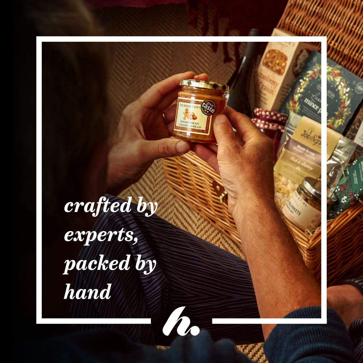 An informative graphic showing a person holding a small jar of gingerbread and caramel spread in front of a wicker basket filled with packaged food items. The scene is warmly lit and includes the text 'crafted by experts, packed by hand' with a logo at the bottom.