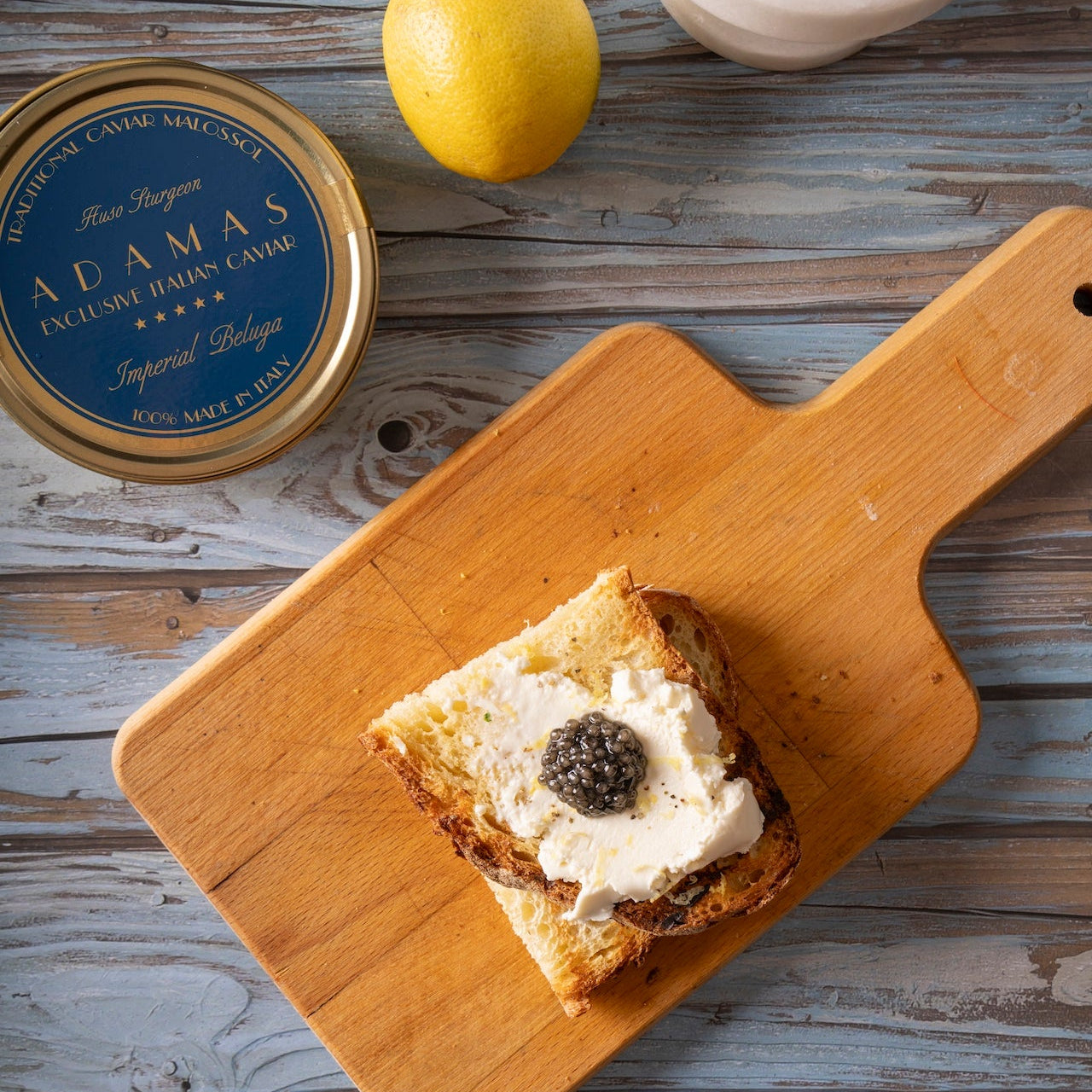 A styled food scene on a rustic weathered wood surface showing a slice of toast topped with white cream and dark caviar, placed on a wooden cutting board. A round tin of Adamas Caviar sits alongside, with a lemon, and a white mortar and pestle.