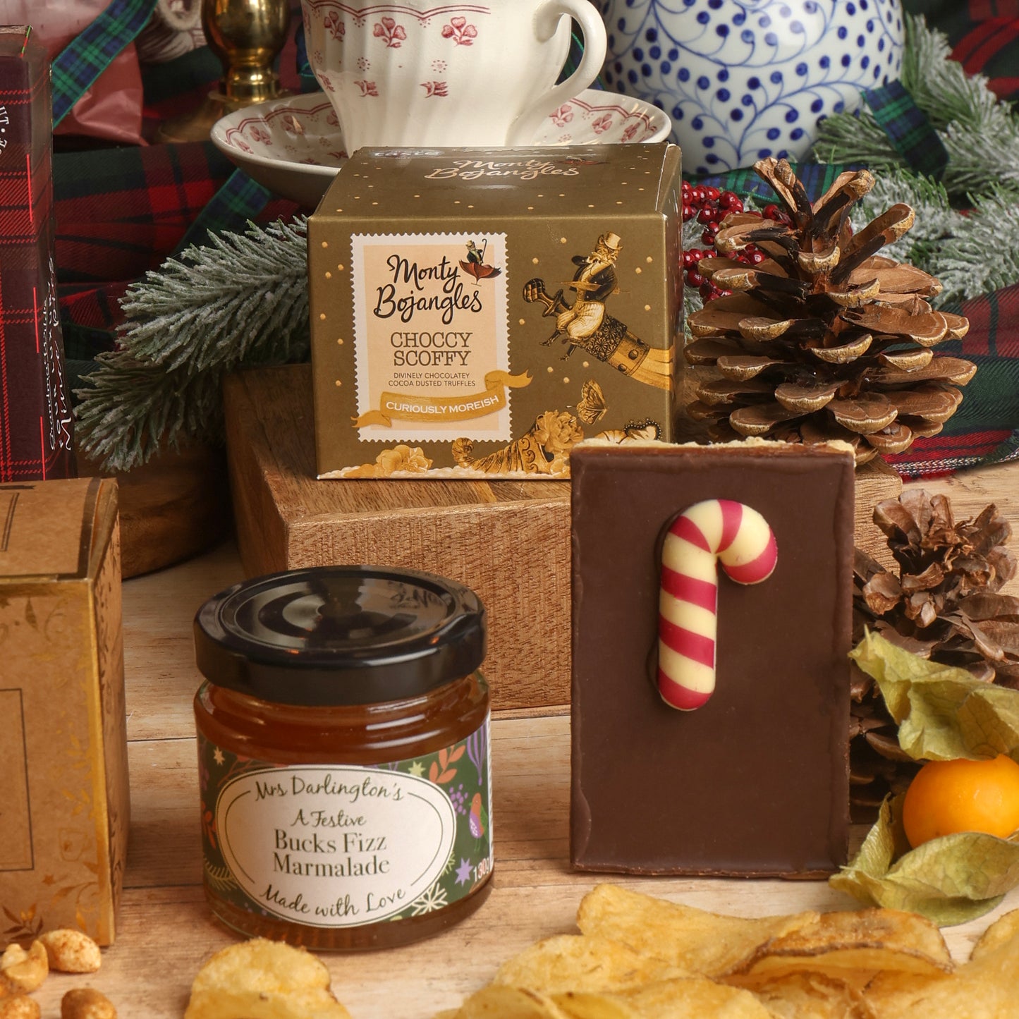 A styled Christmas gift scene showing contents of the Festive Favourites Santa Sack Hamper, including a jar of Bucks Fizz Marmalade, a chocolate flapjack with a red and white candy cane decoration, and boxed chocolate truffles by Monty Bojangles on a wooden surface with crisps, pinecones, and seasonal decorations in the background.