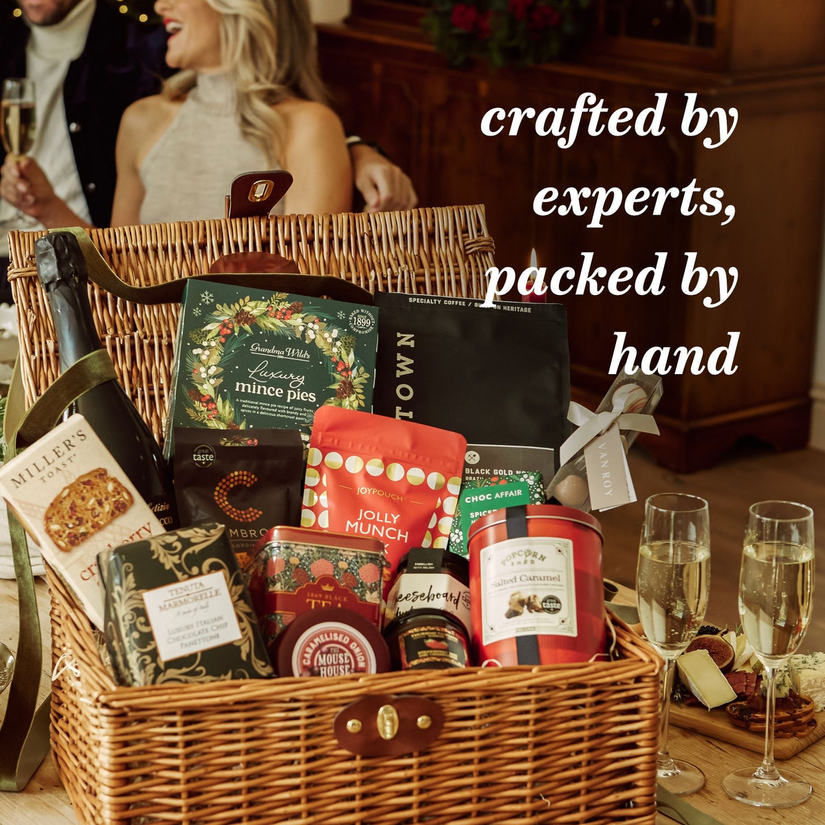 An informative USP image showing a gift basket arrangement with packaged snacks and treats including mince pies, cheese and panettone. White text on the image reads 'crafted by experts, packed by hand'.