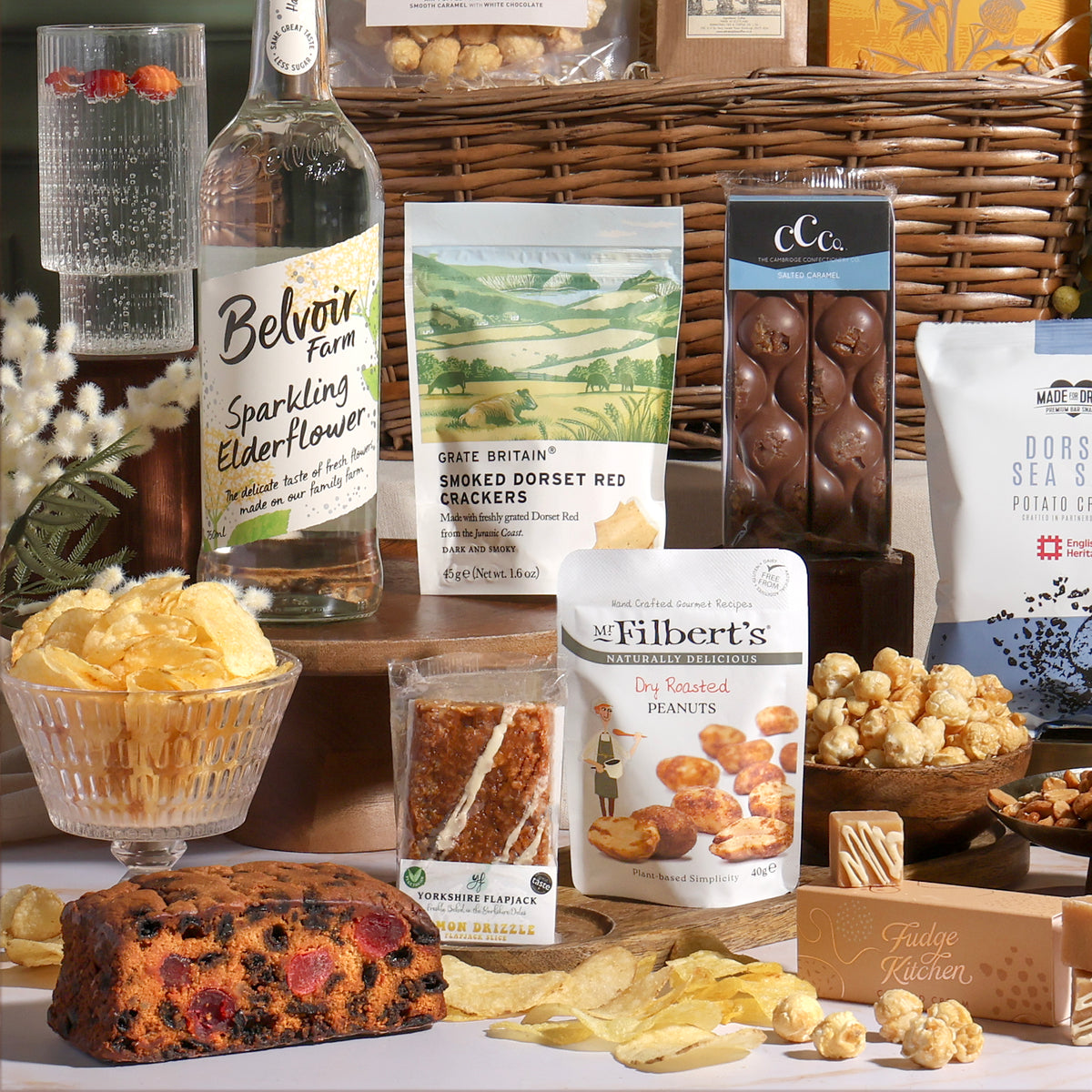 Classic alcohol-free gift basket, with sweet and savoury treats arranged around the willow basket, including salted caramel chocolate, nuts, flapjack, cake, fudge, and sparkling elderflower drink.
