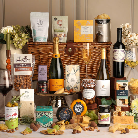 Deluxe Food and Wine Hamper arranged in a wicker basket with Champagne, wine, chocolates, cheeses, nuts, and tea on a table, styled with flowers and wine glasses against a beige panelled wall.