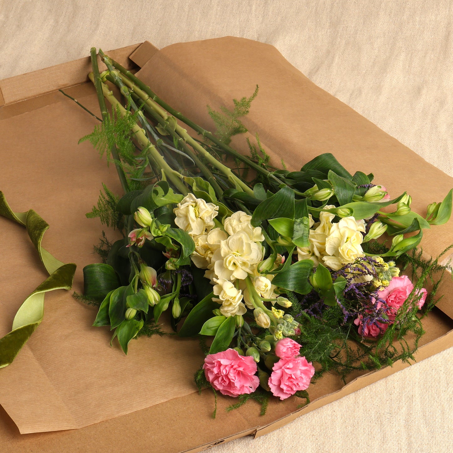 A letterbox bouquet delivery wrapped in brown gift paper inside a brown gift box, featuring mixed seasonal flowers, displayed on a beige fabric surface.