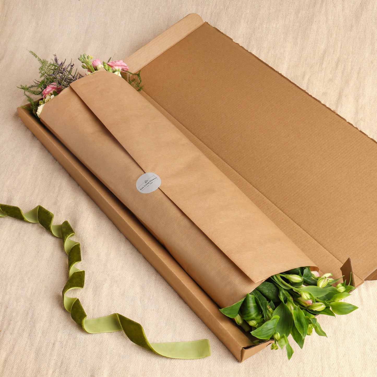 Letterbox flower bouquet wrapped in brown gift paper and presented in a matching brown gift box, with foliage visible at the opening and a green ribbon on a light beige fabric background.