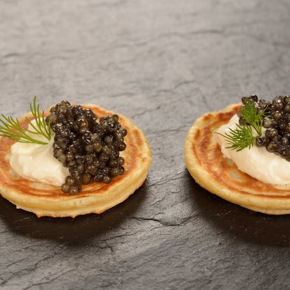 Three small blinis topped with cream and caviar, each garnished with a sprig of dill, arranged on a dark slate surface.
