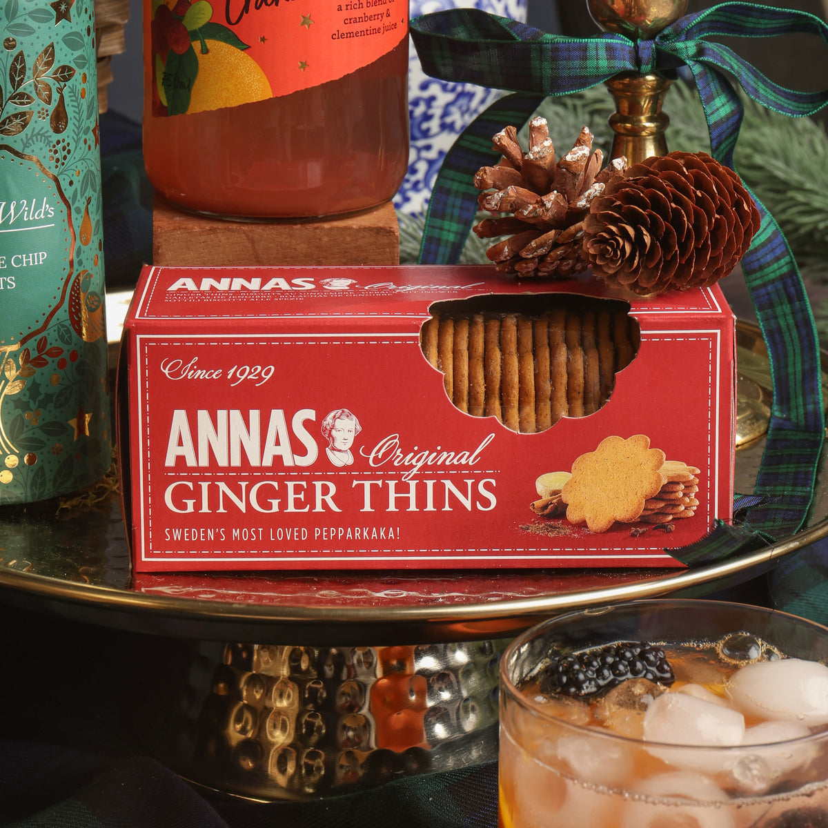 A red box of Anna's Original Ginger Thins biscuits sits on a decorative tray, surrounded by Christmas props including pinecones, a plaid ribbon, and a drink in the foreground.