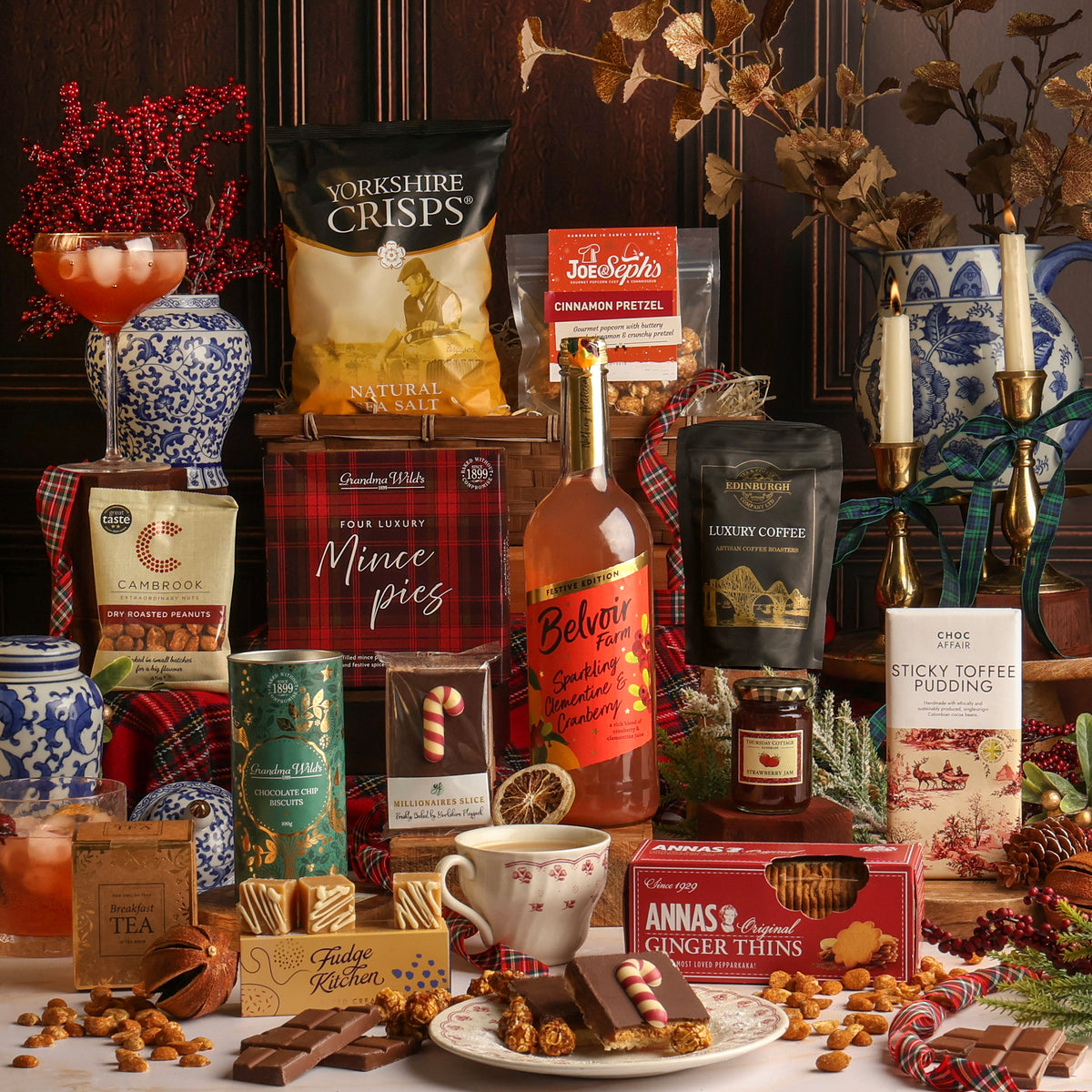 Luxury Joy of Christmas Hamper filled with assorted treats and snacks, including Yorkshire Crisps, sparkling clementine and cranberry, coffee, and Anna's Ginger Thins, arranged with mugs, chocolate, and festive decorations on a table.