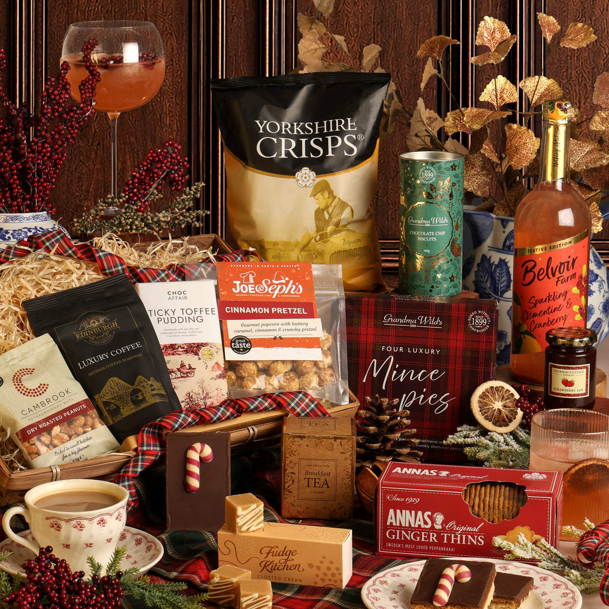 The Luxury Joy of Christmas gift hamper filled with assorted British treats and snacks, including Yorkshire Crisps, Cinnamon Pretzel popcorn, Anna's Ginger Thins, mince pies, and tea, arranged on a plaid table runner with a cup of tea, wine glass with soft drink, and festive decorations.