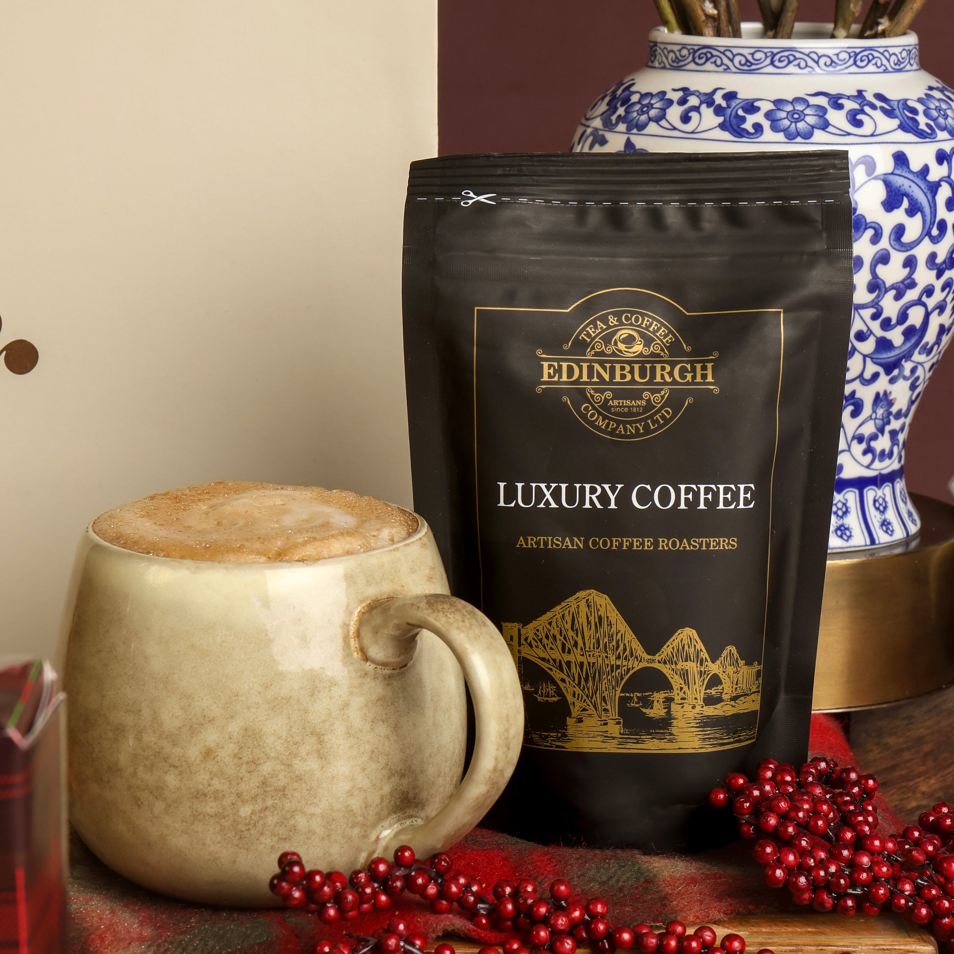 A bag of Edinburgh Luxury Coffee stands on a tabletop beside a ceramic mug filled with coffee, with red berry garlands, and a blue and white decorative vase in the background.