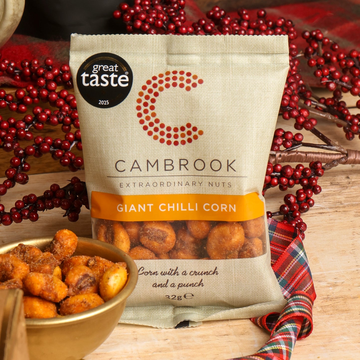 A bag of Cambrook Giant Chilli Corn from the Little Taste of Christmas Hamper with the corn displayed in a small serving bowl, in front of a red berry garland, on a wooden table.