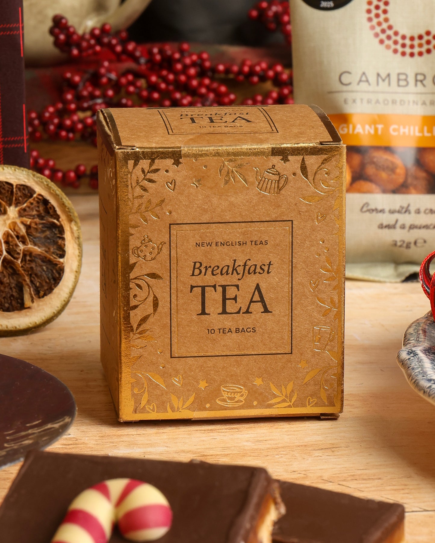 A square box filled with Breakfast Tea stands on a wooden surface, surrounded by tea-related accessories including dried fruit, chocolate flapjack with a candy cane decoration, in a styled tabletop scene.