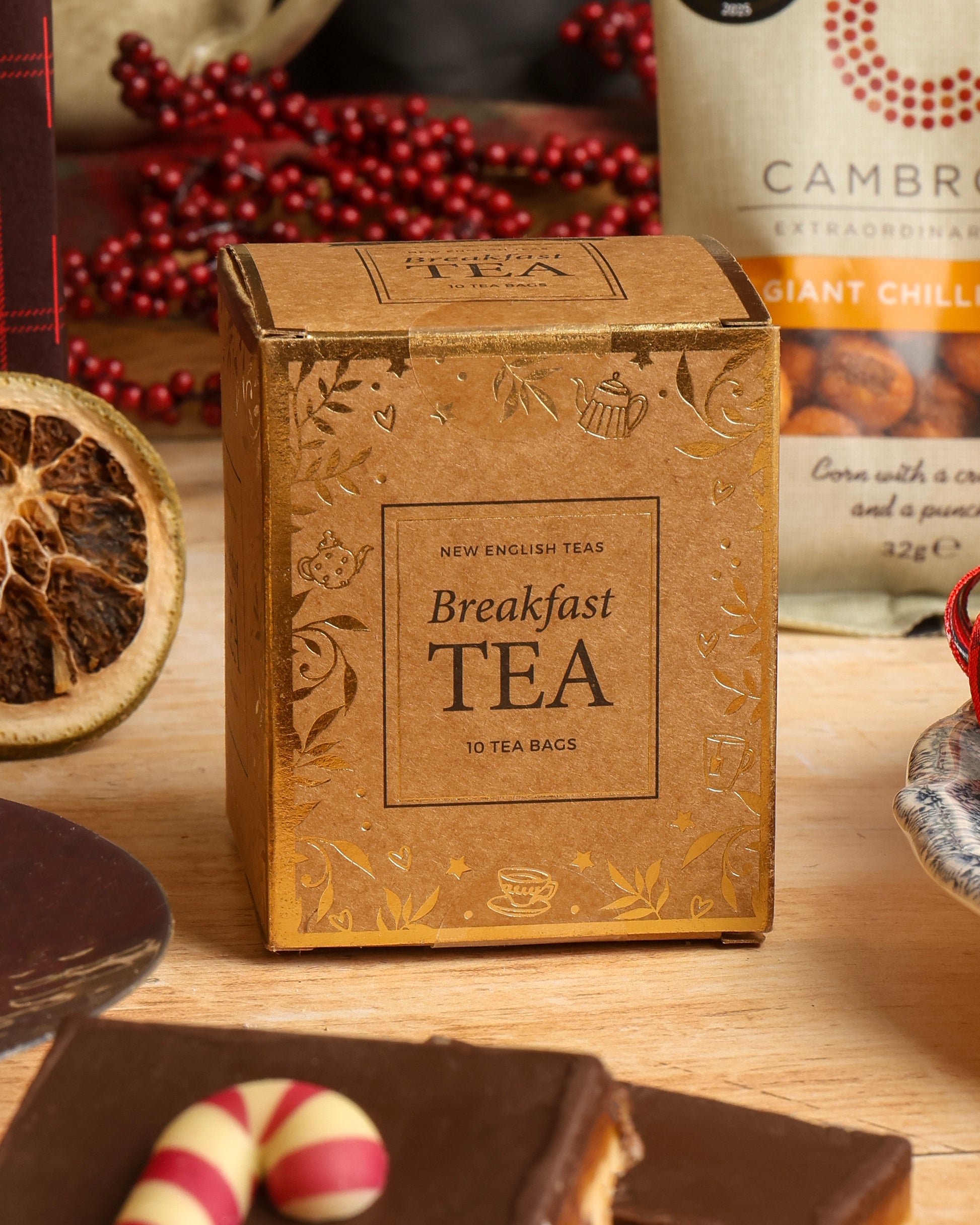 A square box filled with Breakfast Tea stands on a wooden surface, surrounded by tea-related accessories including dried fruit, chocolate flapjack with a candy cane decoration, in a styled tabletop scene.