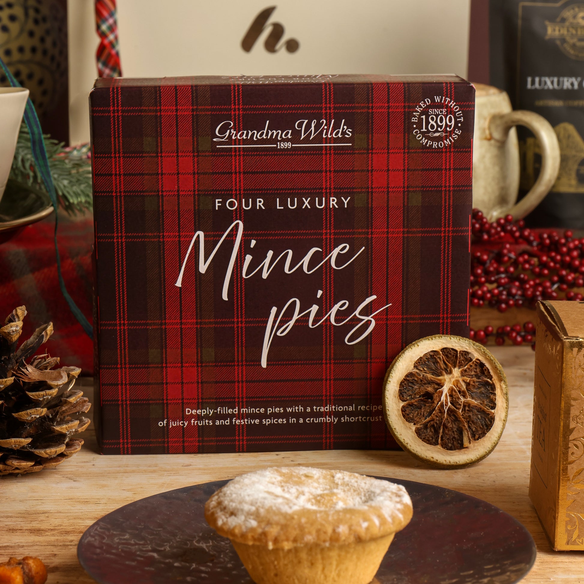 A box of Grandma Wild's Four Luxury Mince Pies sits on a wooden table with a single mince pie and a dried orange slice in front. The box features a red and black plaid pattern with white text.