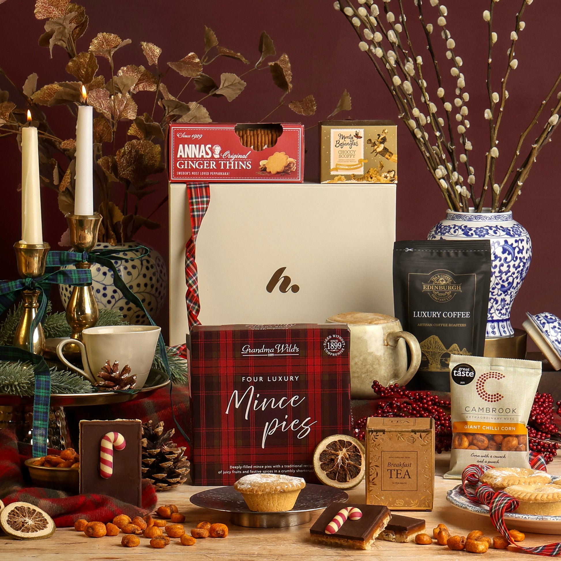 Little Taste of Christmas Hamper with the food contents arranged on a table with candles, a ceramic vase, pine cones, winter floral decorations, and dried citrus slices, against a dark burgundy background with a white branded gift box at the back. The food presented includes chilli corn, a box of mince pies, candy cane flapjack, tea, luxury ground coffee, and chocolate truffles.