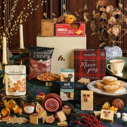 Christmas Season Selection Gift Box hamper with the food and drink contents displayed, featuring crisps, biscuits, tea, cheese, and mince pies, set against a wooden panel background with candles, festive foliage, and a plate of mince pies.