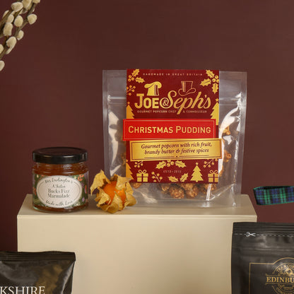 A cream-colour gift box stands with a bag of Joe and Seph's Christmas Pudding popcorn on top, along with a jar of marmalade and a dried orange decoration, against a dark brown background.