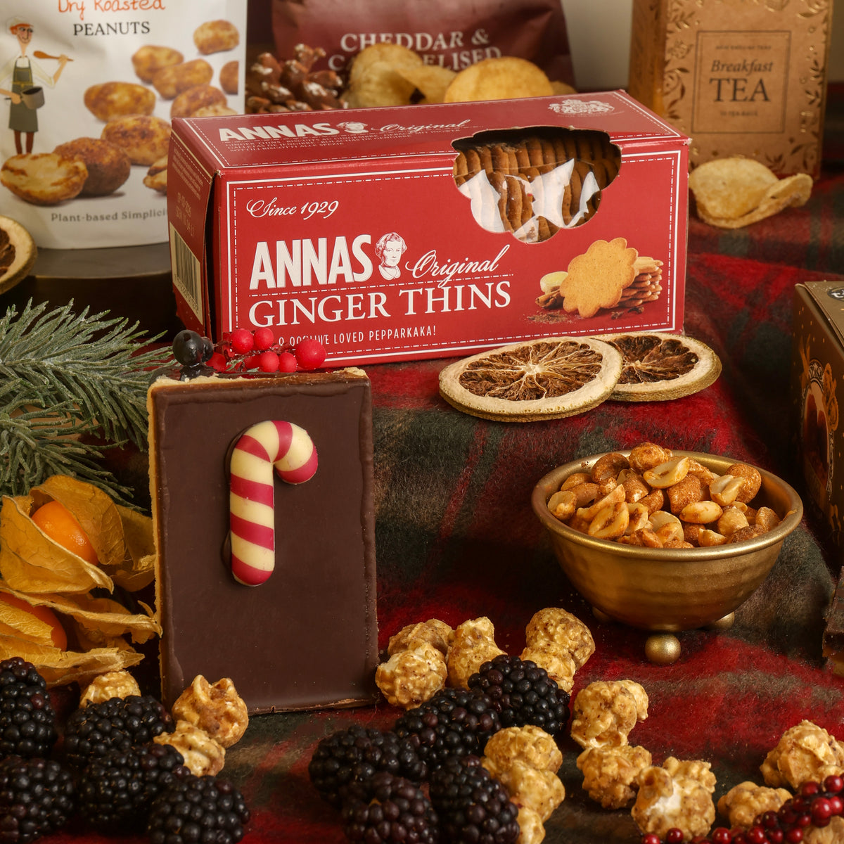 A styled Christmas spread featuring a red box of Anna's Original Ginger Thins, a chocolate flapjack with a candy cane decoration, a bowl of nuts, dried citrus slices, blackberries, popcorn, and pine branches on a red plaid cloth.