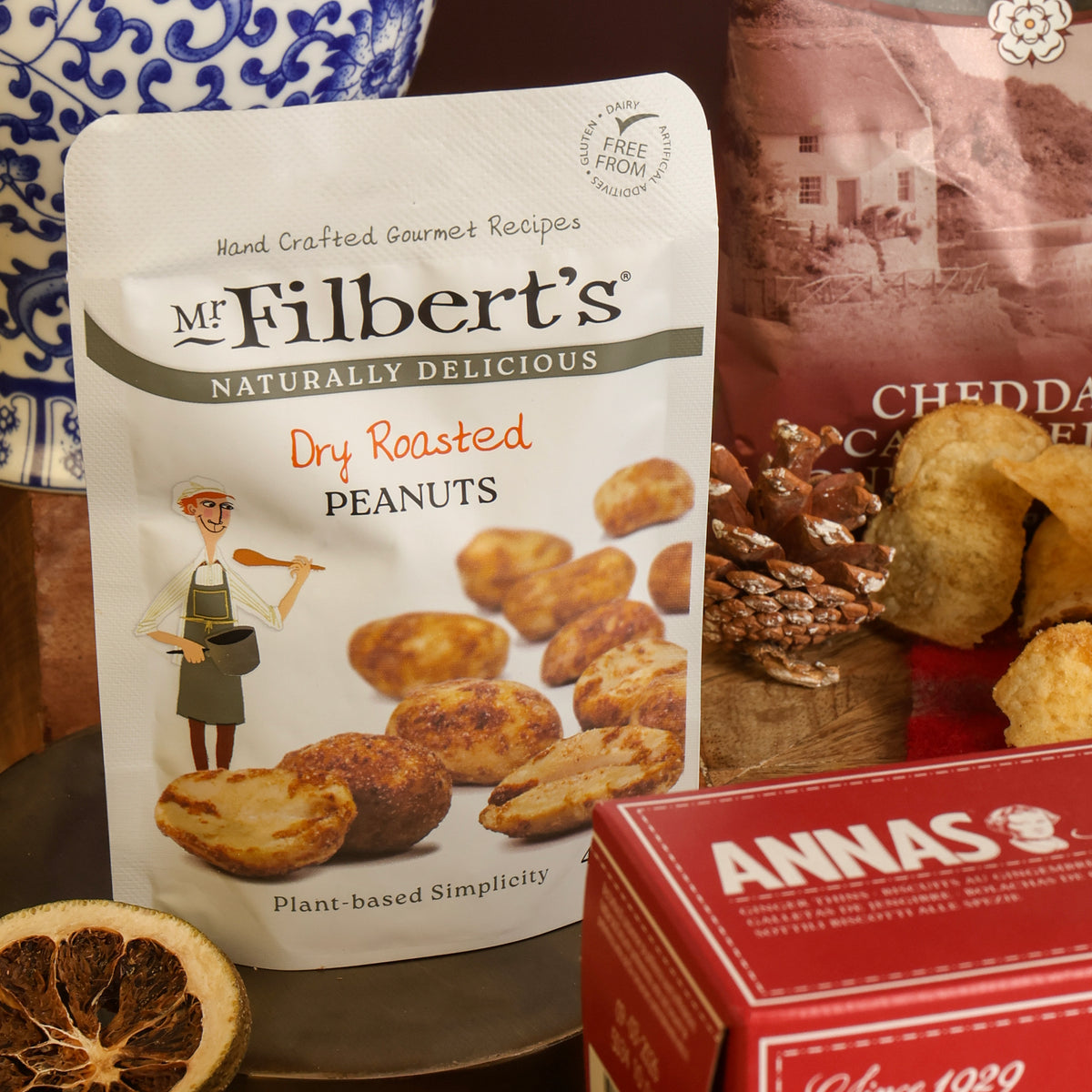 A stand-up pouch of Mr. Filbert's Dry Roasted Peanuts in the front of a snack assortment display, with a red box of biscuits, a blue-and-white ceramic pot, and crisps on a tabletop.