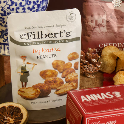 A stand-up pouch of Mr. Filbert's Dry Roasted Peanuts in the front of a snack assortment display, with a red box of biscuits, a blue-and-white ceramic pot, and crisps on a tabletop.