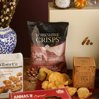 A styled snack and tea assortment on a tabletop, featuring a bag of Yorkshire Crisps in the centre, with crisps scattered in front. Surrounding products include a pouch of Mr. Filbert's dried roasted peanuts, a red box of biscuits, and a brown tea box.
