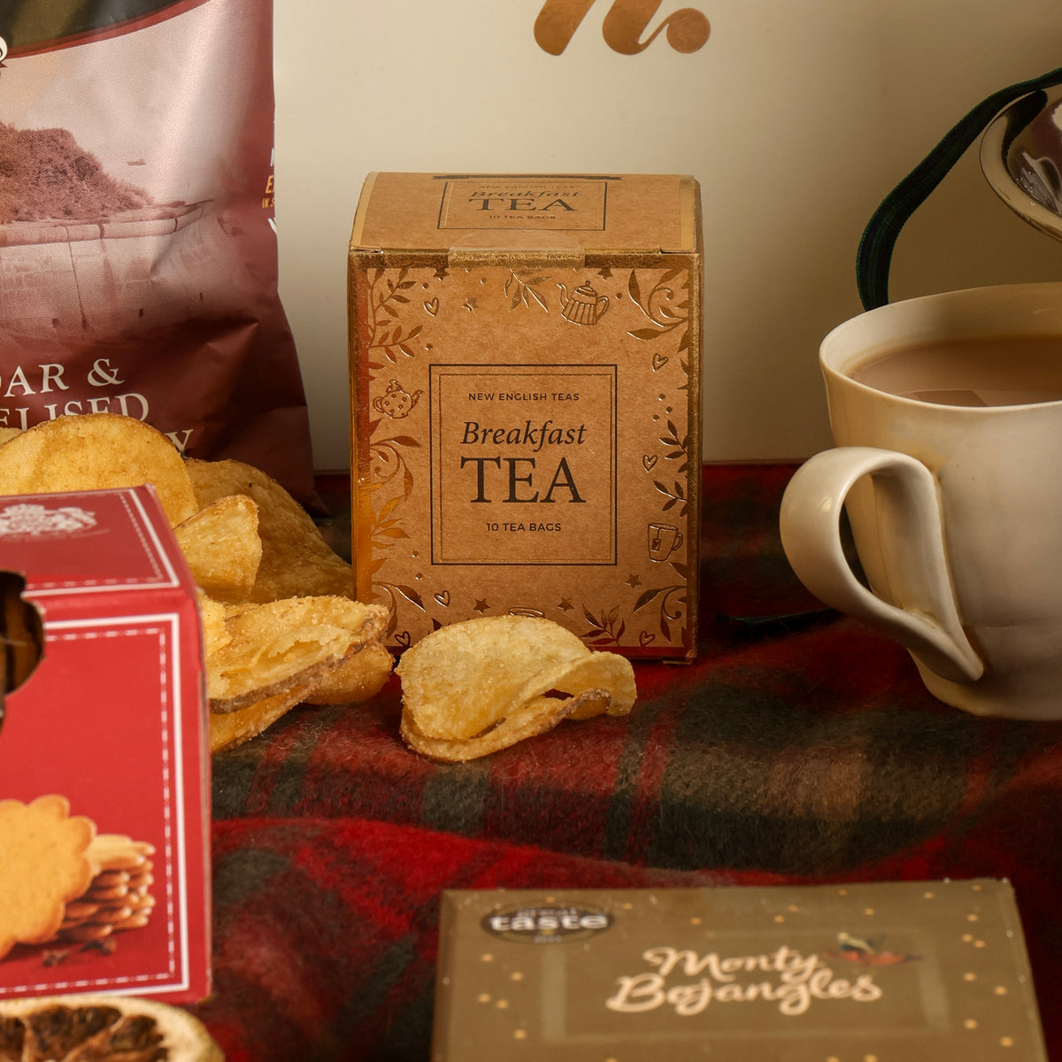 A styled tabletop scene featuring a box of Breakfast Tea in the centre, with a white mug of tea on the right, biscuits on the left, and other packaged snacks around it on a red plaid cloth.
