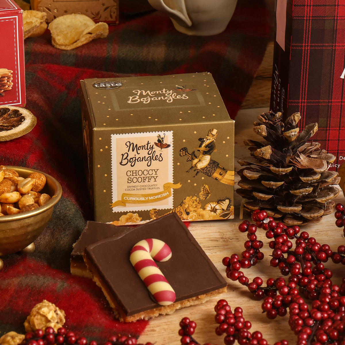 A box of Monty Bojangles chocolate truffles is styled on a wooden surface with a chocolate flapjack, red berries, a pinecone, and Christmas-themed props in a warm, festive setting.