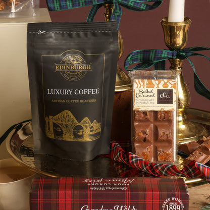 A gift-style product arrangement featuring a black stand-up pouch of Edinburgh Luxury Coffee with gold branding, a Salted Caramel Chocolate Bar, and a red plaid box in the foreground, styled on a tray with a candle and ribbon.