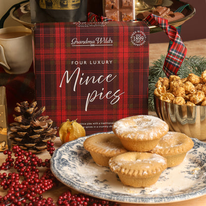 A box of Grandma Wild's Four Luxury Mince Pies sits on a tabletop surrounded by festive Christmas props, including a plate of four mince pies dusted with powdered sugar, a bowl of caramelised popcorn, red berries, pinecones, and a Christmas ribbon.