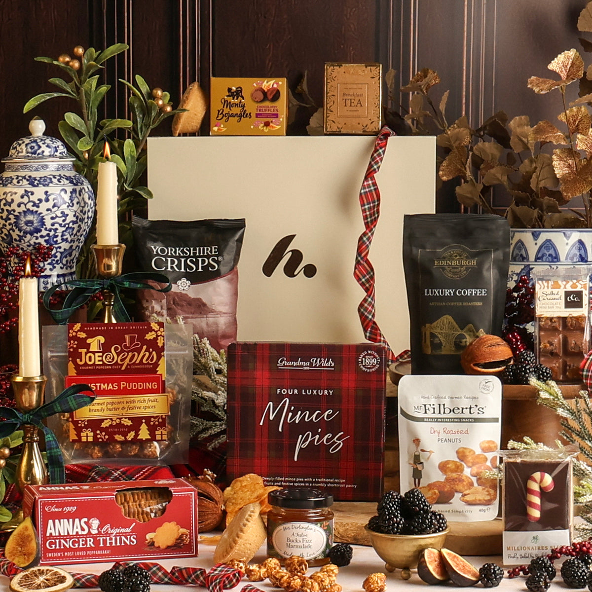 A Christmas-themed Bearing Gifts hamper featuring an assortment of British snacks, sweets, and coffee arranged around a central cream-colored gift box with a red plaid ribbon.