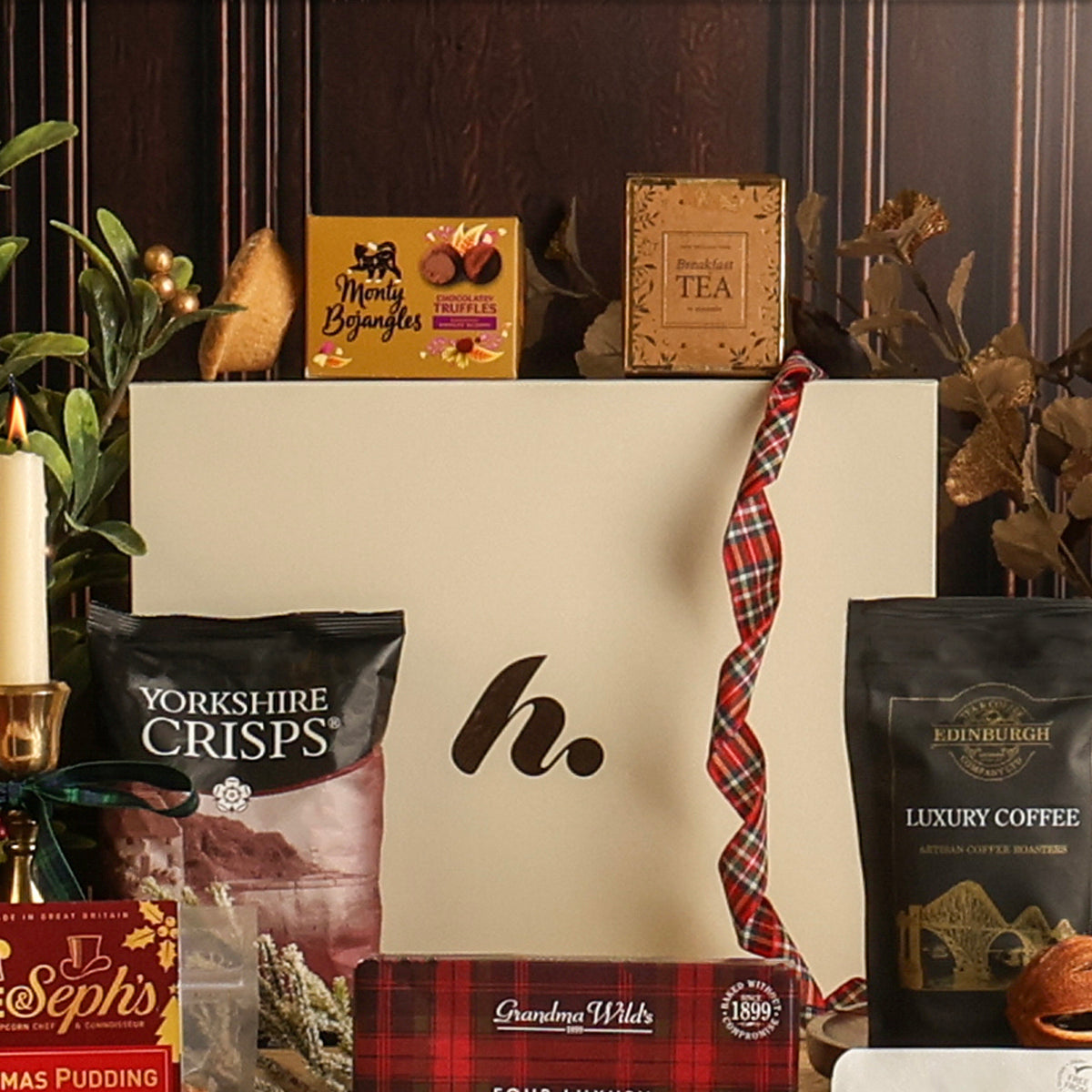 Bearing Gifts Christmas gift hamper, displaying a curated arrangement featuring a large cream-coloured gift box, surrounded by assorted packaged treats and coffee, styled with a red plaid ribbon against a dark wood background.