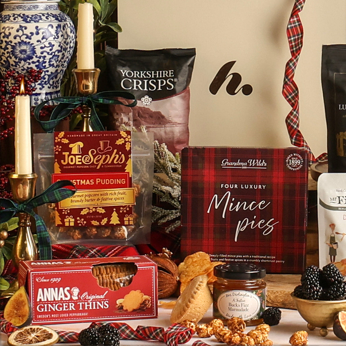 A styled Bearing Gifts Christmas gift hamper with assorted British treats and snacks, including Yorkshire Crisps, Joe & Seph's Christmas Pudding popcorn, Anna's Original Ginger Thins, and mince pies, arranged with candles, blackberries, and plaid ribbon.