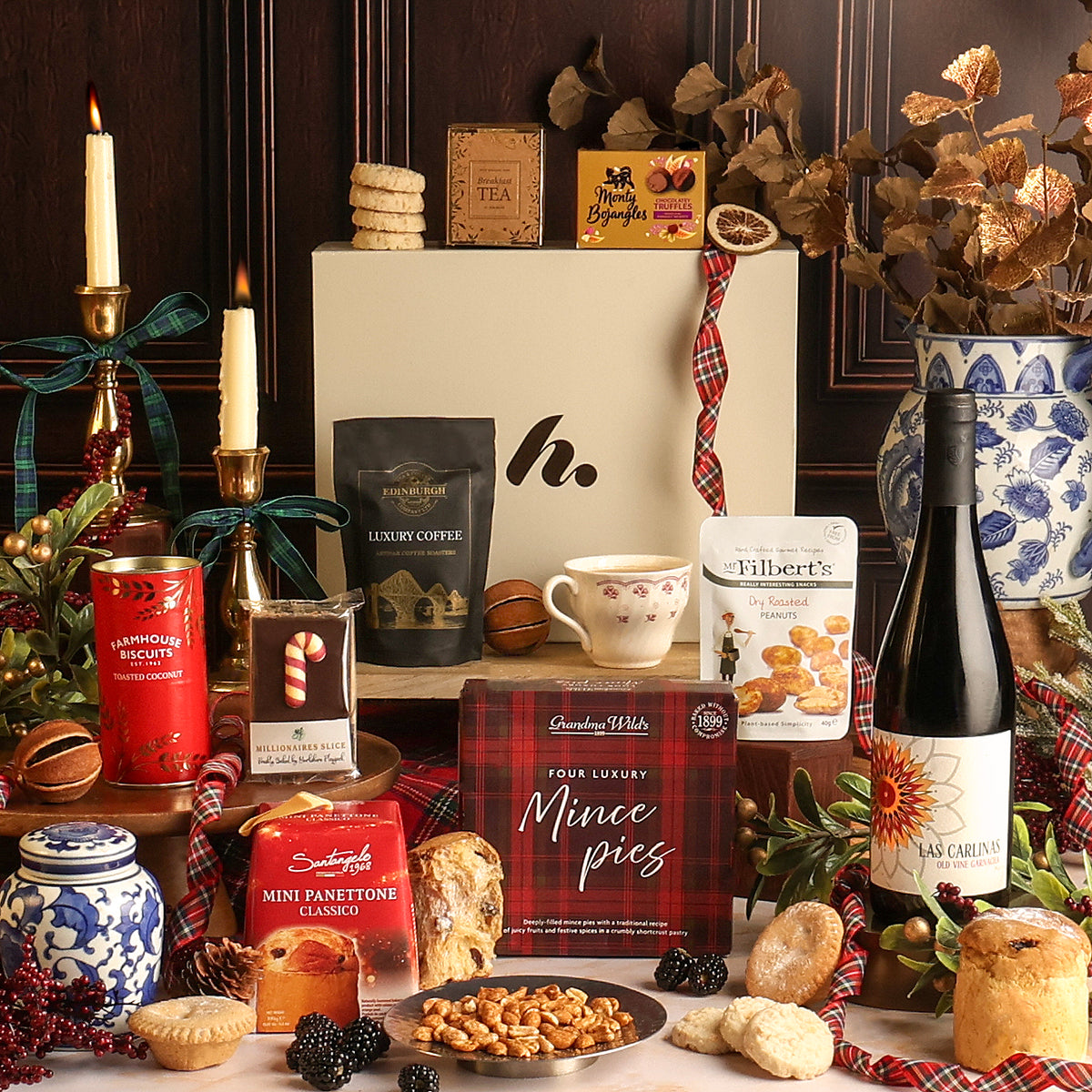 The Christmas Cracker Hamper arranged on a table with candles, ceramic jars, dried foliage, and assorted food items including mince pies, biscuits, tea, coffee, and a bottle of wine. A cream gift box with a red plaid ribbon stands in the centre back.