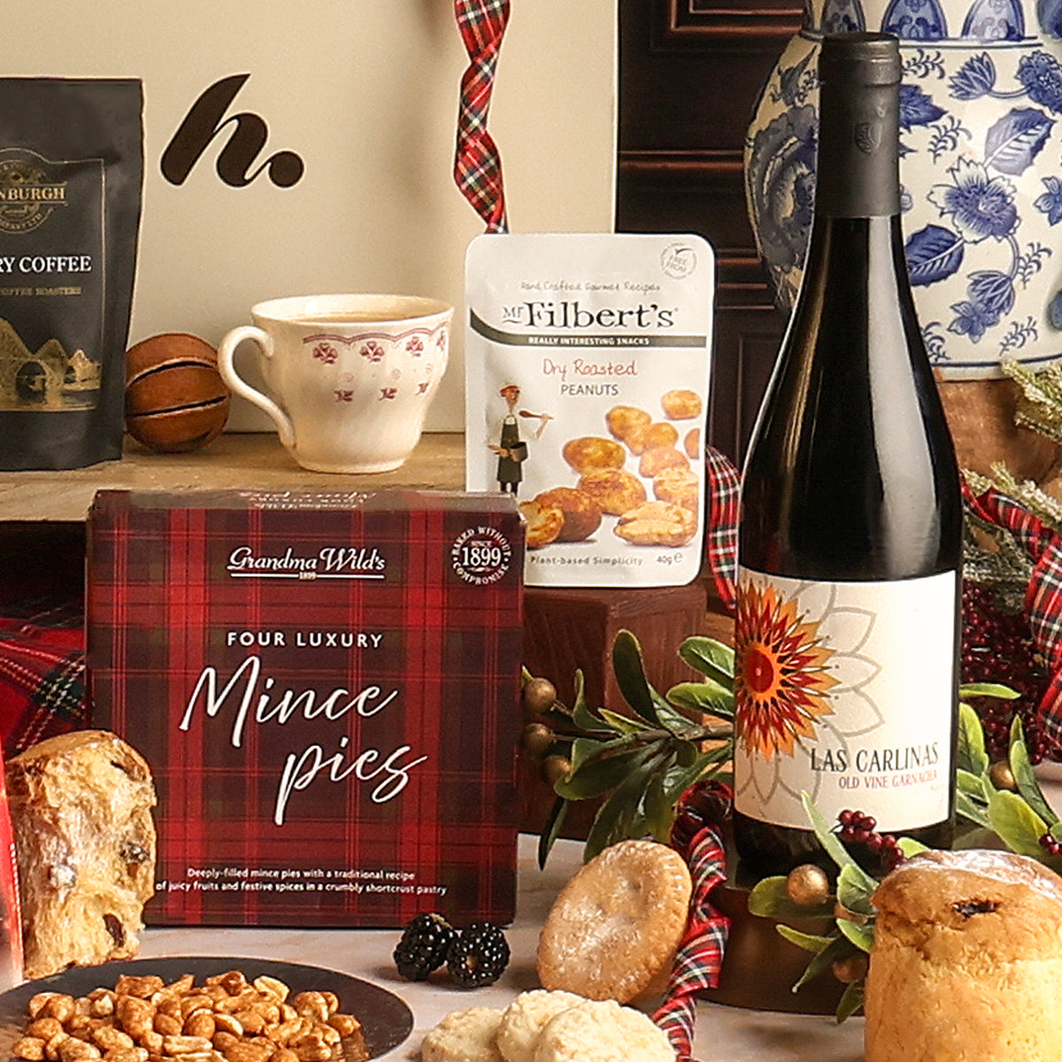 A Christmas-themed gift basket arrangement featuring a box of Grandma Wild's Four Luxury Mince Pies, a bag of Mr. Filbert's Dry Roasted Peanuts, a bottle of Las Carlinas red wine, a cup of tea, with assorted biscuits and nuts on a rustic wooden surface.
