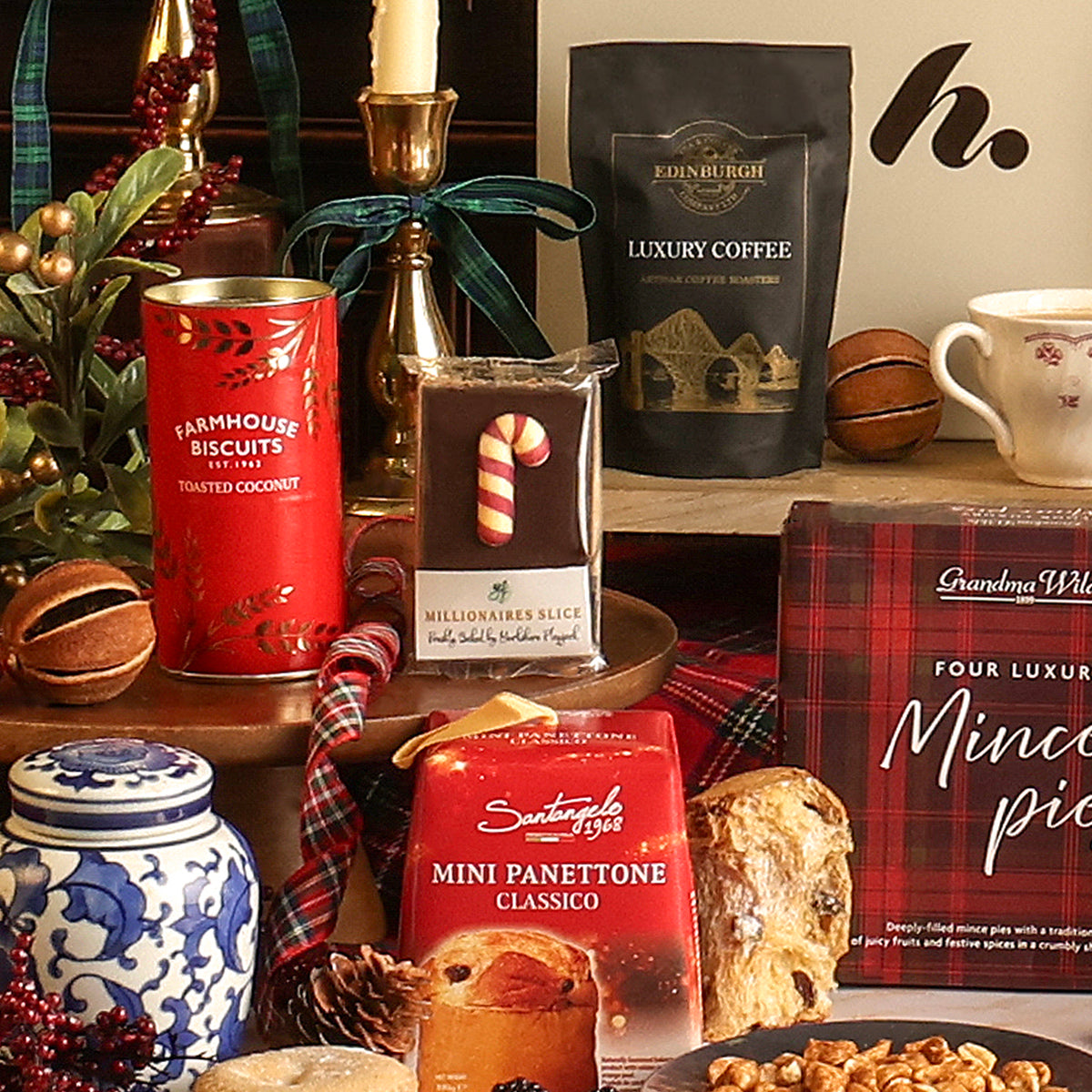 The Christmas Cracker gift hamper filled with assorted gourmet treats and coffee, including a red tin of Farmhouse Biscuits toasted coconut biscuits, a bag of Edinburgh Luxury Coffee, a millionaire's slice, a mini panettone, and a mince pie box, styled with seasonal decor on a wooden surface.