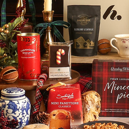 The Christmas Cracker gift hamper filled with assorted gourmet treats and coffee, including a red tin of Farmhouse Biscuits toasted coconut biscuits, a bag of Edinburgh Luxury Coffee, a millionaire's slice, a mini panettone, and a mince pie box, styled with seasonal decor on a wooden surface.