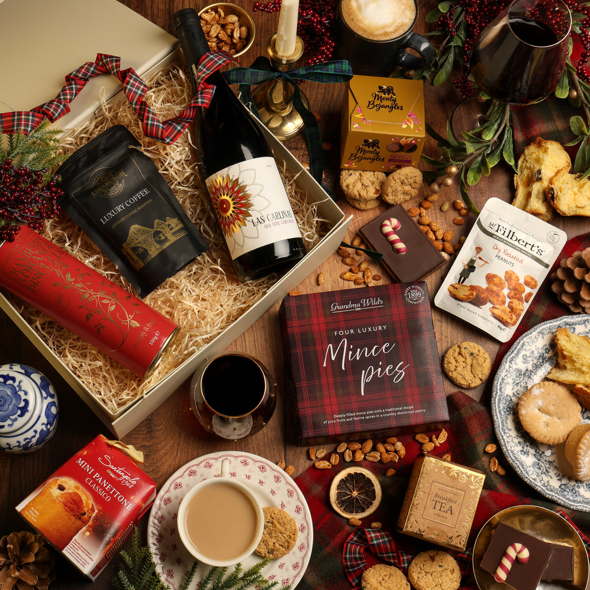 A festive gift hamper styled in a cosy Christmas flat lay on a wooden table. The cream-colour gift box filled with shredded paper contains a bottle of red wine, coffee and biscuits, surrounded by a box of mince pies, chocolate flapjack, a mug of tea, and festive decorations.
