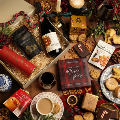 A festive gift hamper styled in a cosy Christmas flat lay on a wooden table. The cream-colour gift box filled with shredded paper contains a bottle of red wine, coffee and biscuits, surrounded by a box of mince pies, chocolate flapjack, a mug of tea, and festive decorations.