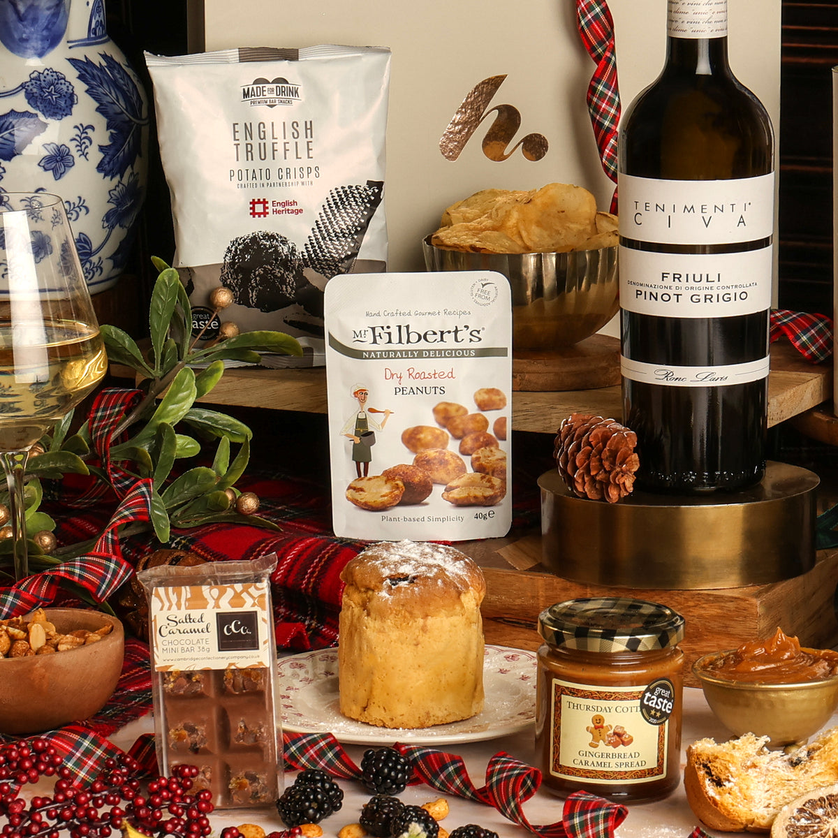 A styled festive food and drink spread featuring a bottle of Pinot Grigio white wine, a bag of Mr. Filbert's peanuts, English Truffle potato crisps, a Salted Caramel chocolate bar, a glass of white wine, a plate with a panettone, and plaid accents on a wooden table.