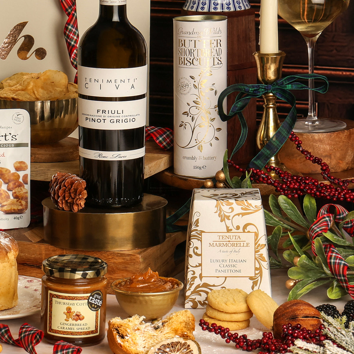 A styled Christmas food and wine spread showing the contents of the Let It Snow hamper, featuring a bottle of Pinot Grigio white wine, a tin of Butter Shortbread Biscuits, a bag of Tenuta Marmorelle panettone, a jar of gingerbread caramel spread, and biscuits, with a glass of white wine on a decorated table.