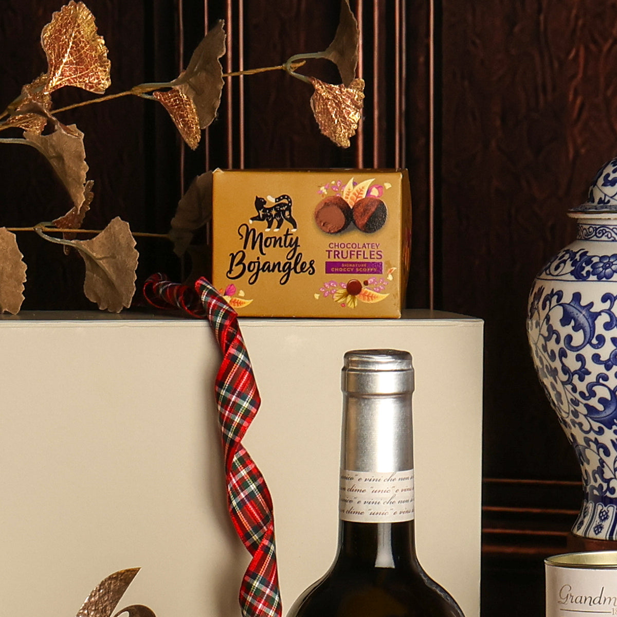 A box of Monty Bojangles chocolate truffles displayed on a white gift box next to a bottle of wine, with a red plaid ribbon, decorative dried leaves, and a blue-and-white ceramic vase in the background.